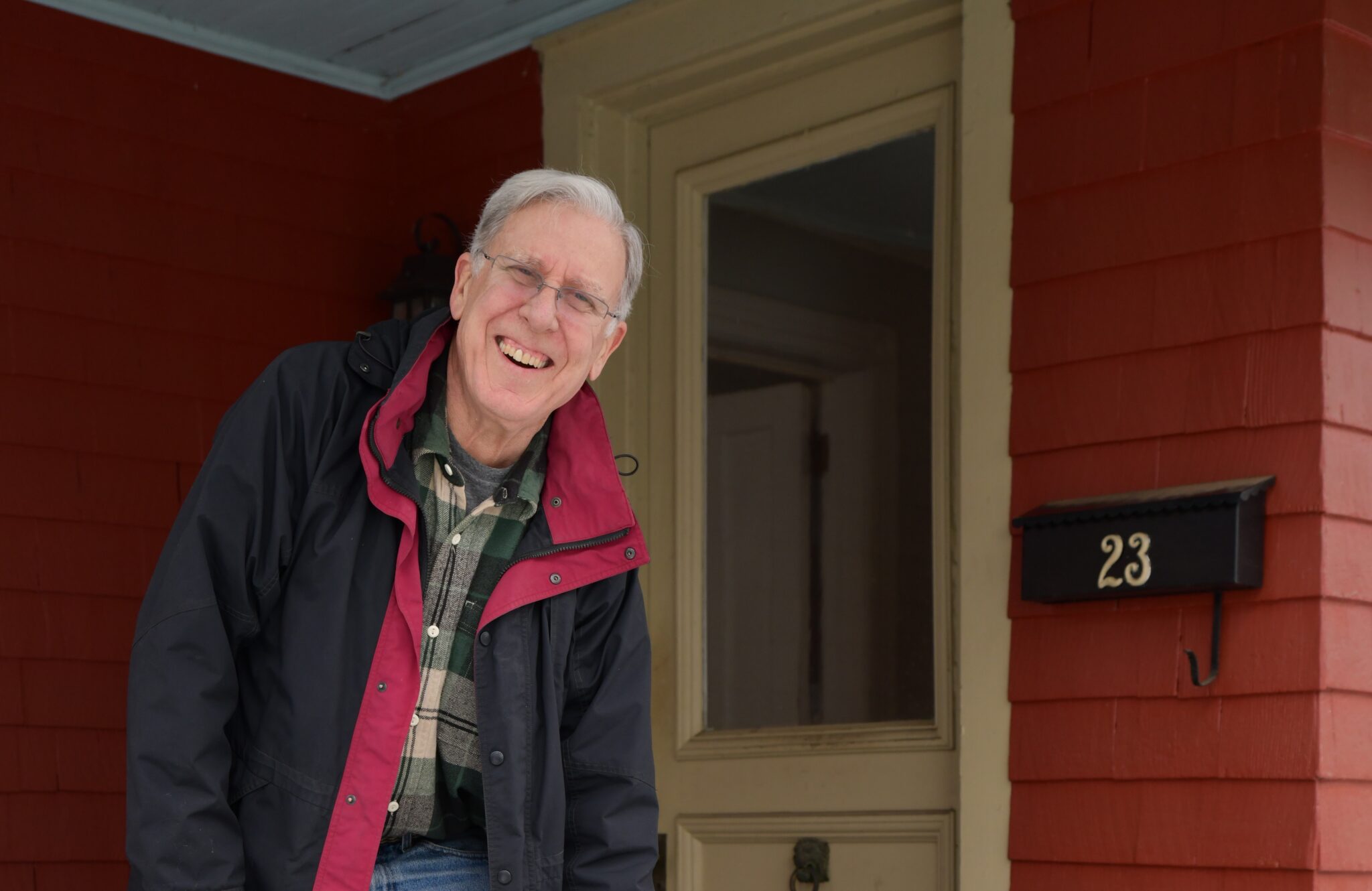 older male with gray hair, glasses, black jacket standing in front of his red house