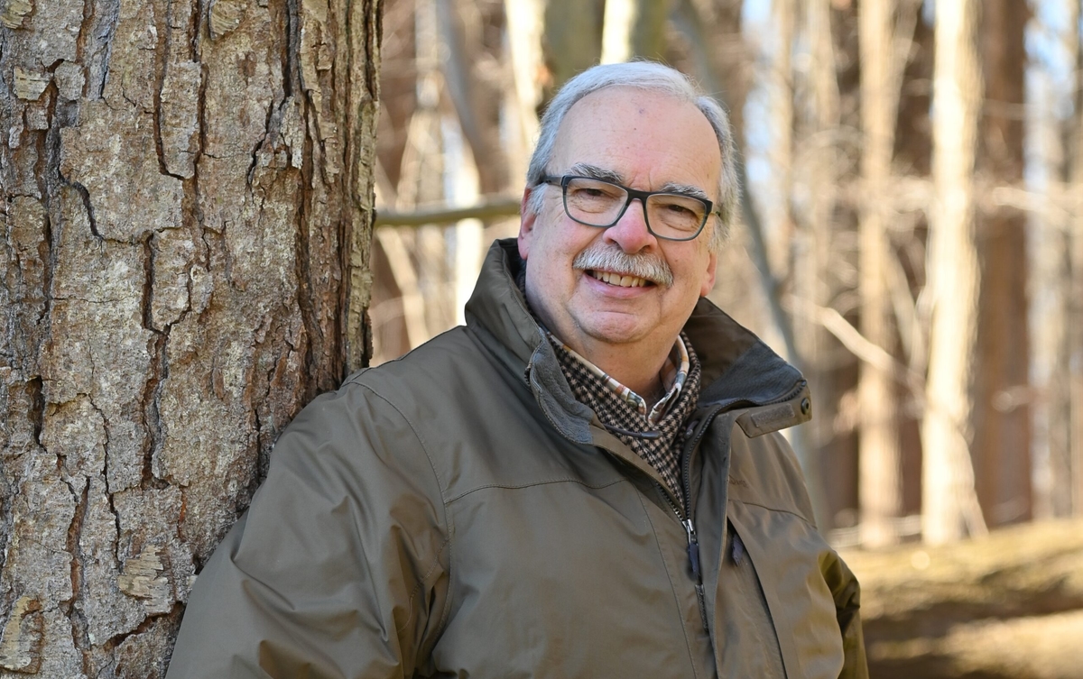 older man with white hair, facial hair and glasses wearing an olive jacket