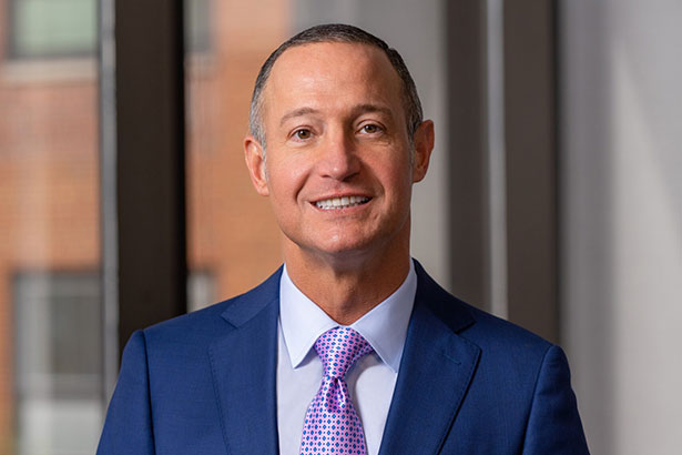 male headshot in blue suit, lavender shirt, and a pink and blue polka dot tie